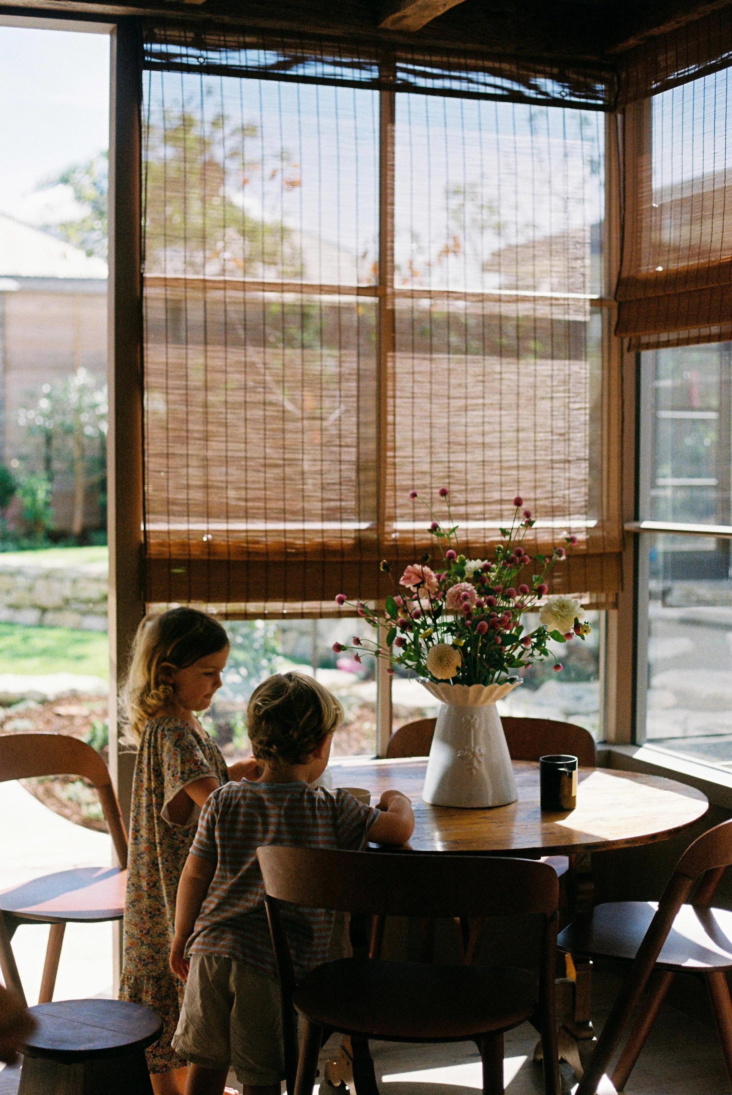 Two children at wooden table with flower vase. Large windows with bamboo blinds. Garden visible outside.