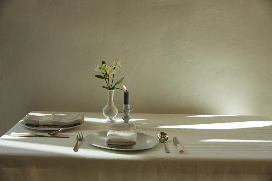 White Belgian Linen tablecloth, vase of flowers, lit candle, plate, cutlery, and folded napkin. Plain wall background, soft lighting.