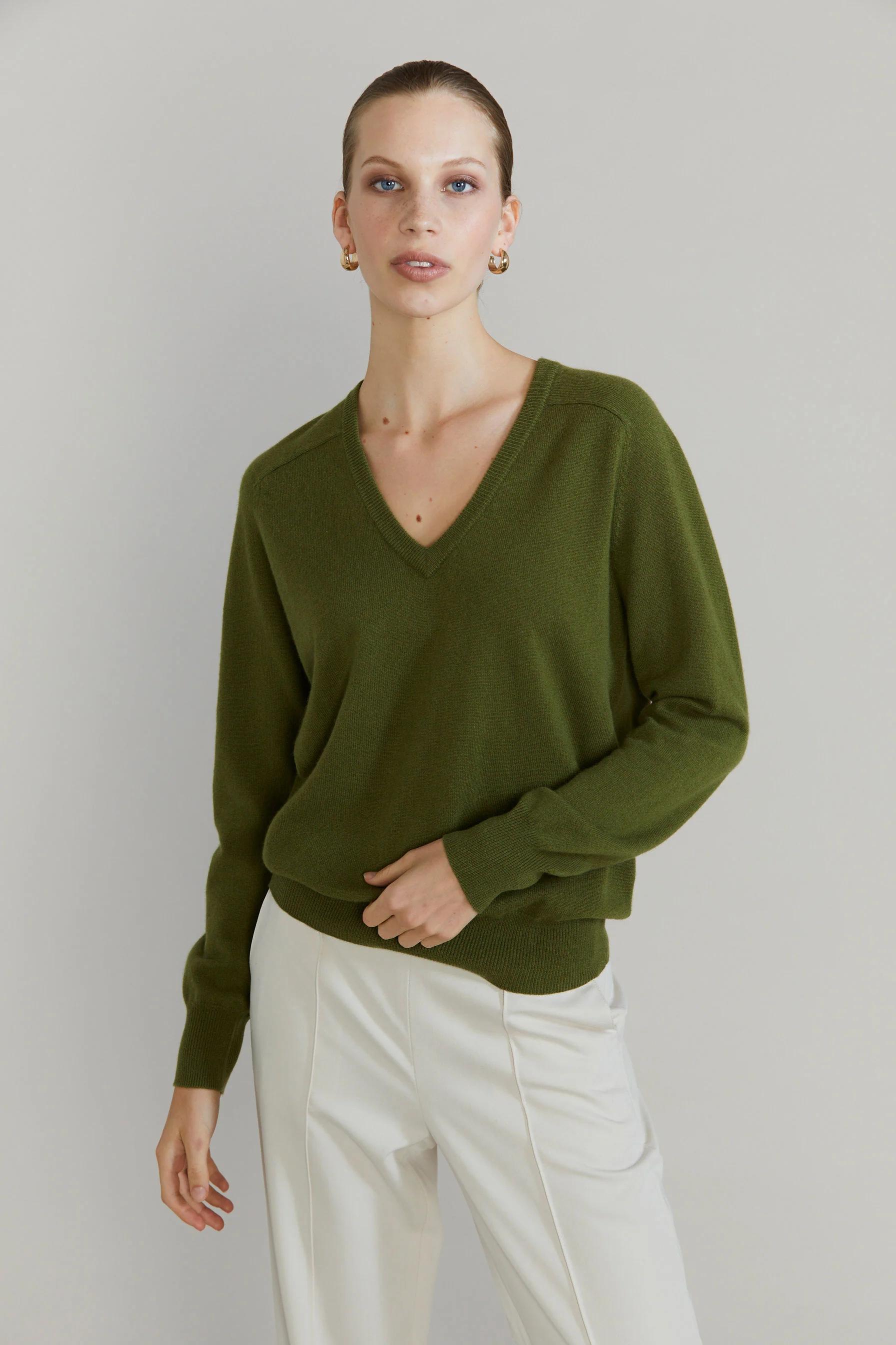 Person in green V-neck sweater and light pants stands against plain background. One hand by side, other near sweater bottom.