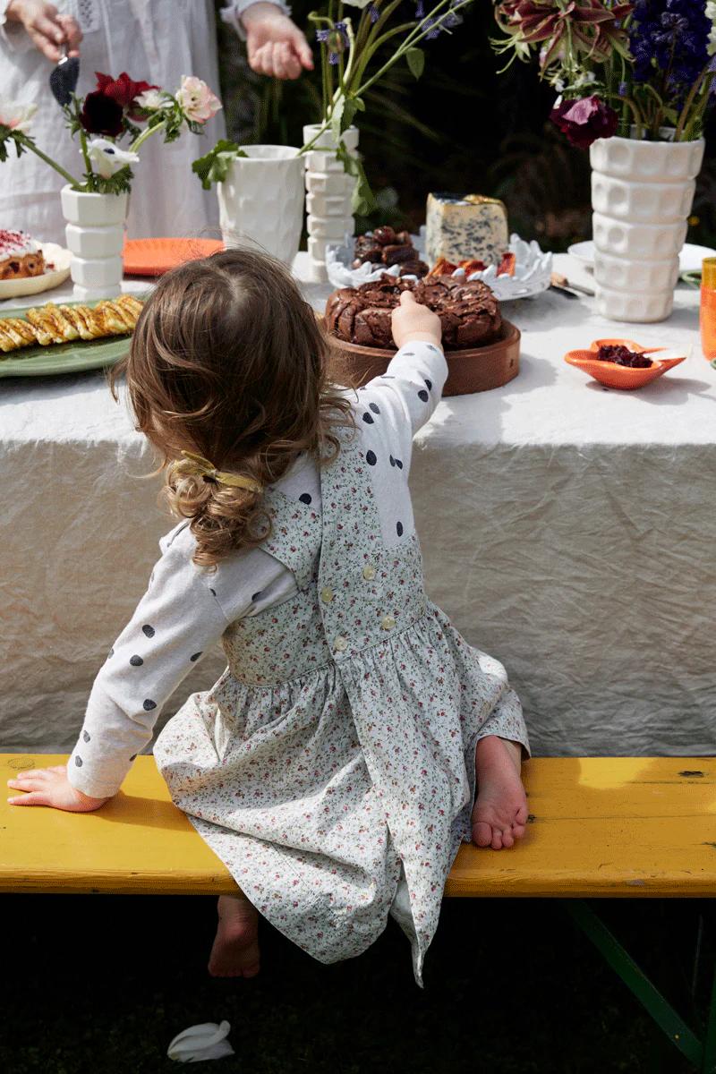 Curly-haired child in floral dress reaches for bundt cake on table filled with food and flowers. Child sits on yellow bench, eyeing baking mix creation.