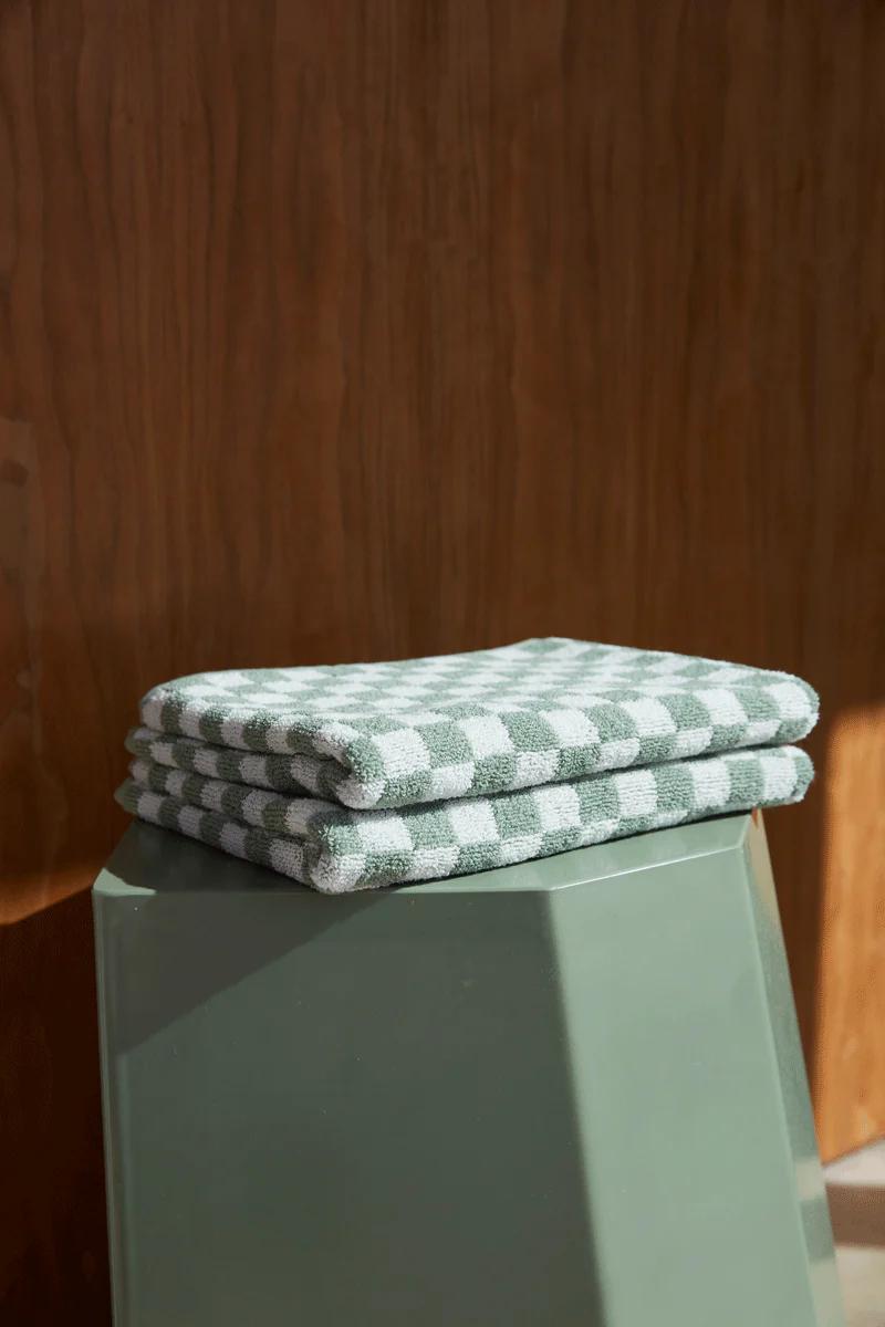 A folded stack of green and white checkered organic cotton towels on a green geometric pedestal. Warm lighting highlights the textures against a wooden surface, showcasing mindful craftsmanship.