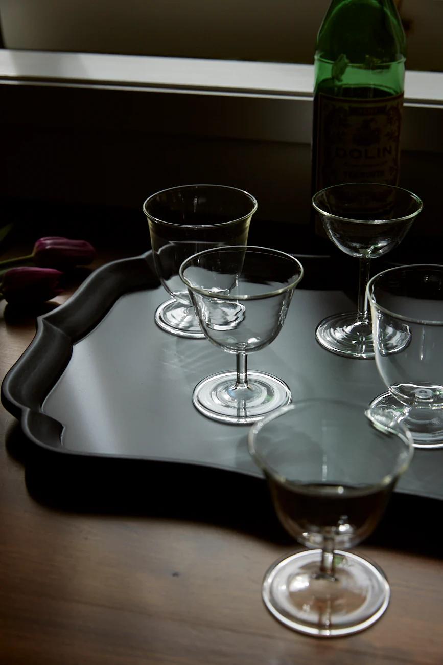 A black tray on a wooden table holds five empty stem glasses. A green bottle stands behind, with blurred flowers in the background.