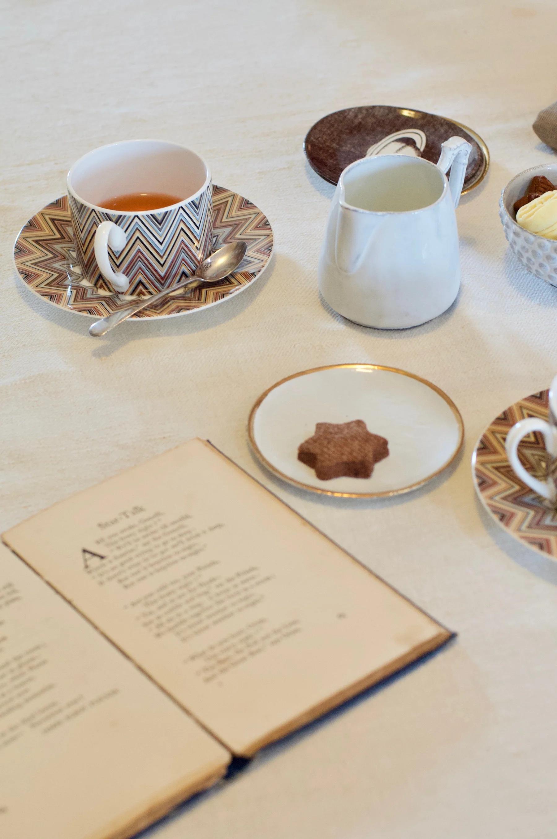 Tea set with zigzag patterned cup and saucer, spoon, jug, plates with cookies, and open book on light tablecloth.