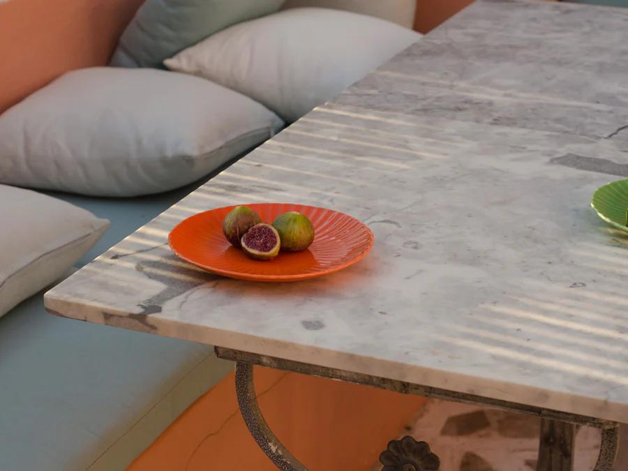 White marble table with orange plate of figs beside L-shaped bench. Pastel pillows and soft shadows create warm garden ambiance.