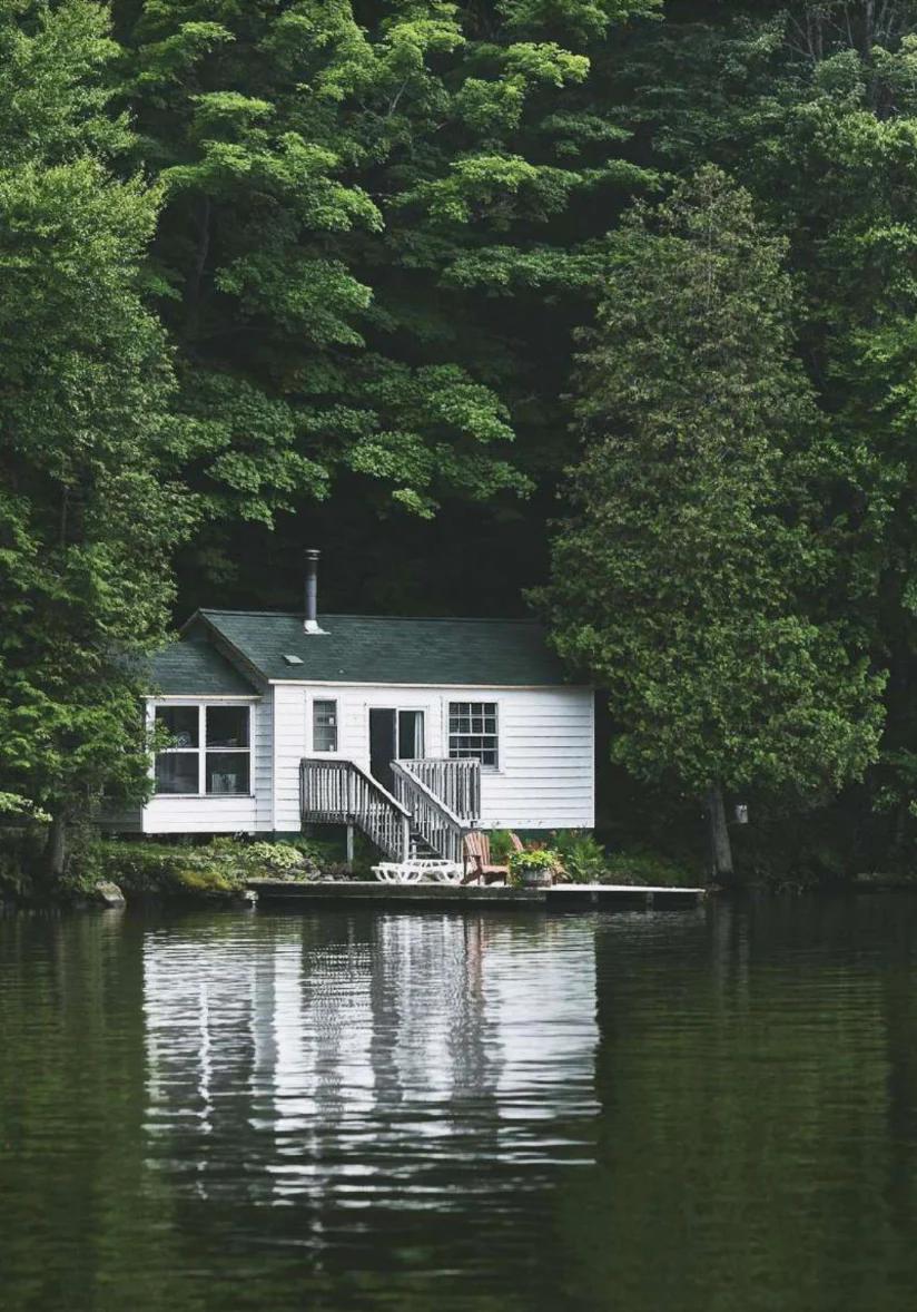 Small white cabin with green trim near calm lake. Wooden deck with railing reflected on water.