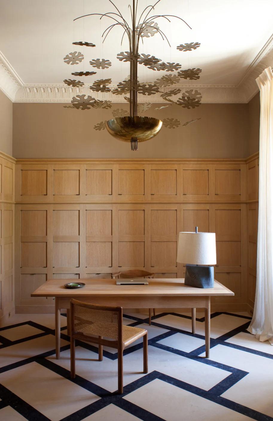 A minimalist château office with wooden desk, stone lamp, and unique chandelier. Beige walls complement the geometric-patterned floor.