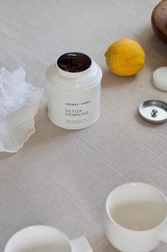 A white canister labeled "Storm + India Detox Morning" on a grey tablecloth with the lid removed. There is also a lemon, cloth napkin, white mug, and the top of a wooden bowl.