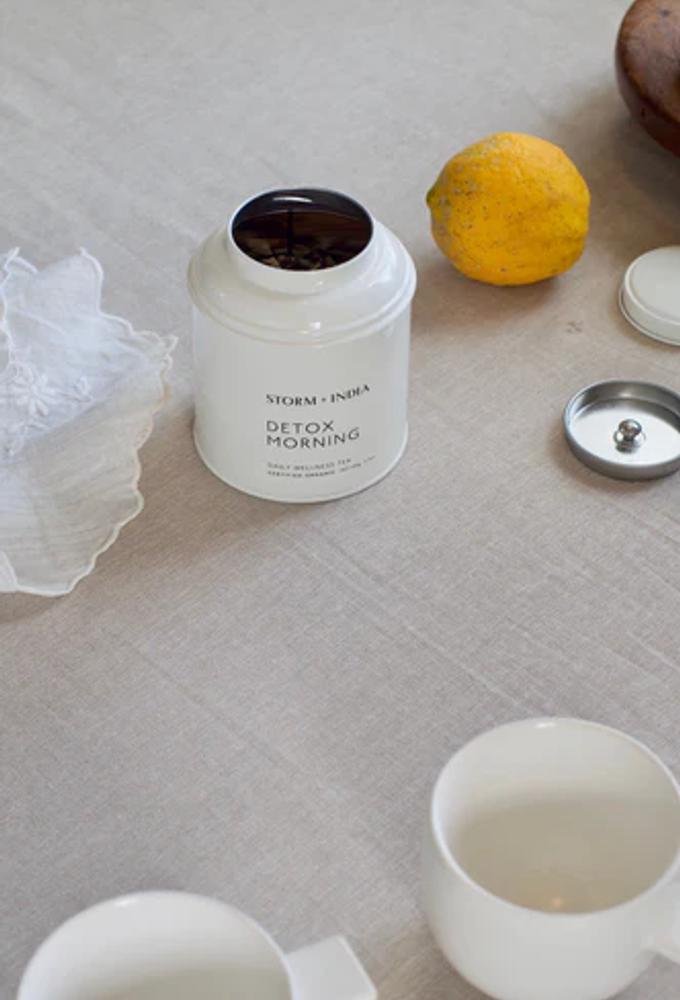 A white canister labeled "Storm + India Detox Morning" on a grey tablecloth with the lid removed. There is also a lemon, cloth napkin, white mug, and the top of a wooden bowl.