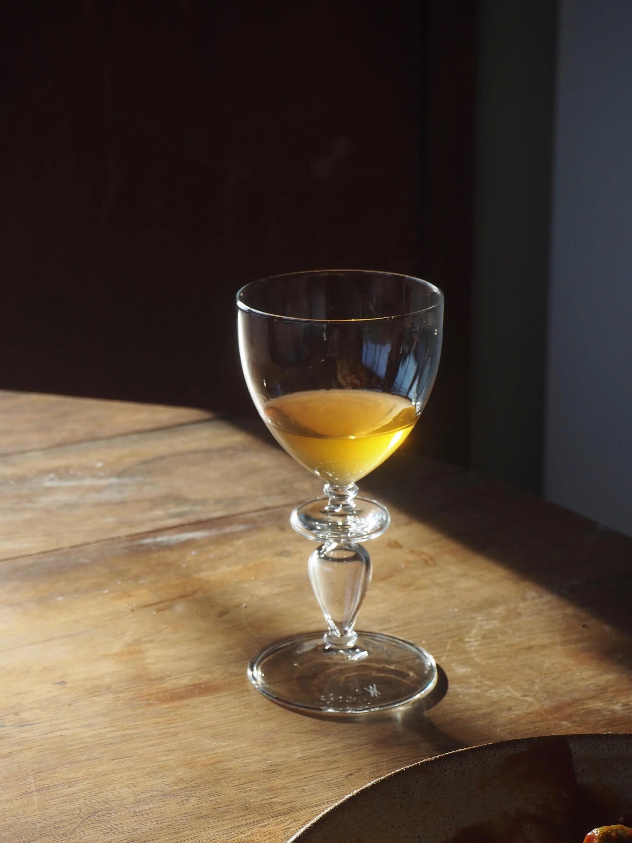 Half-full wine glass with amber organic wine on wooden table. Soft light highlights glass design and casts shadow. Dimly lit background contrasts warm table lighting.