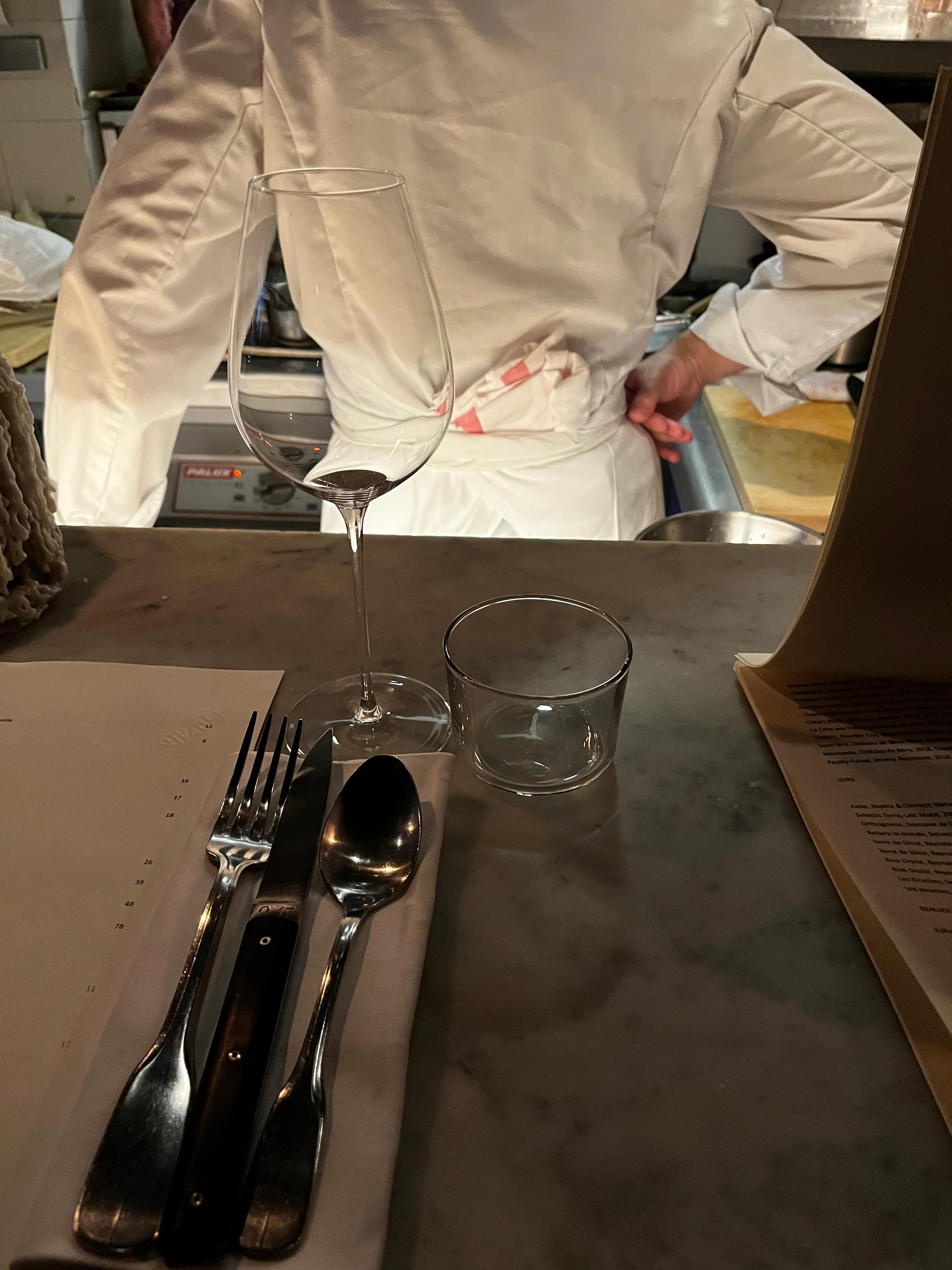 Table setting with glasses, utensils, and napkin on marble surface. Person in white chef's coat with back turned in background.