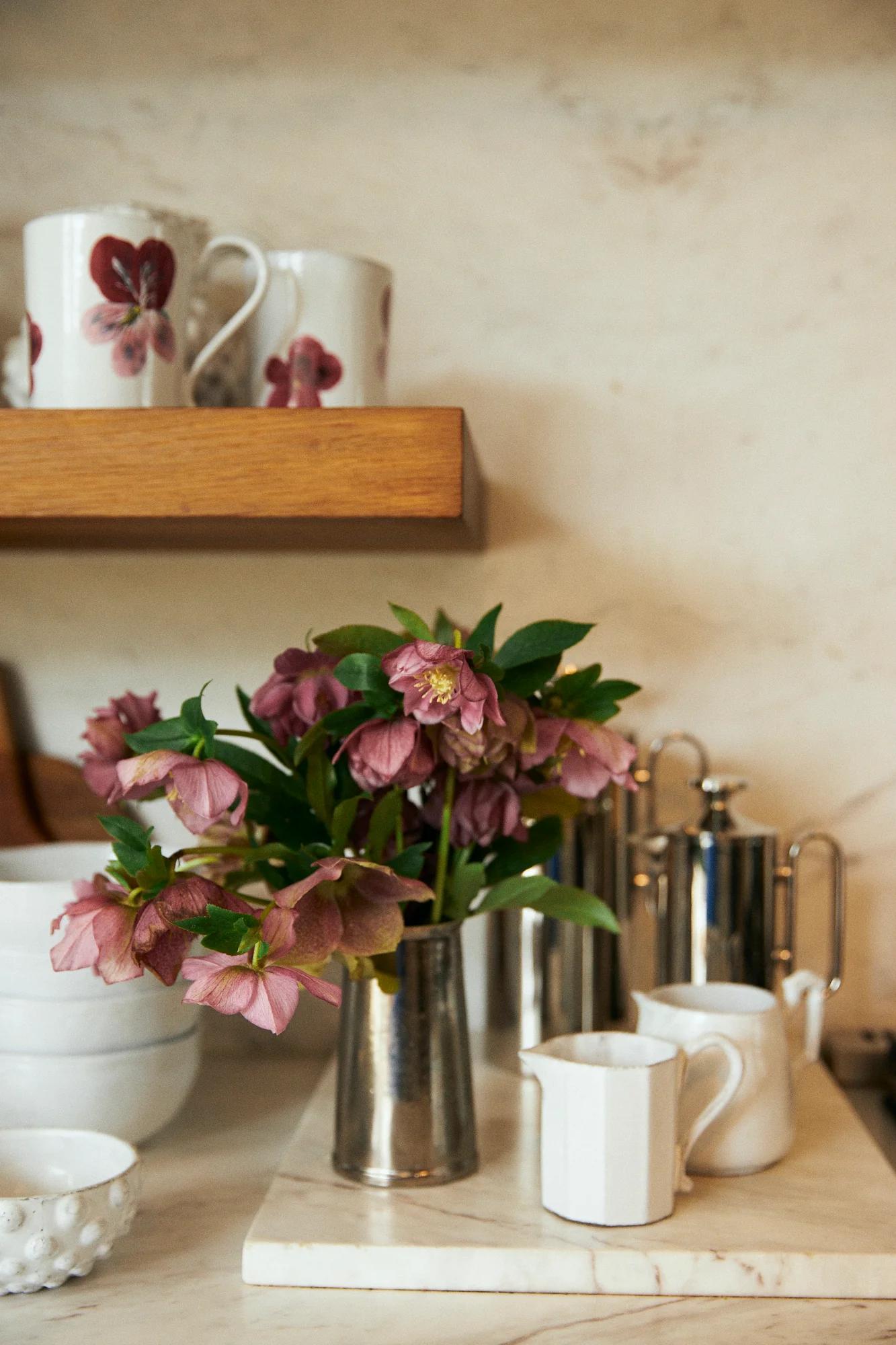 Kitchen counter with flowers in silver vase, sugar bowl, creamer, kettle, and cups with red floral designs.