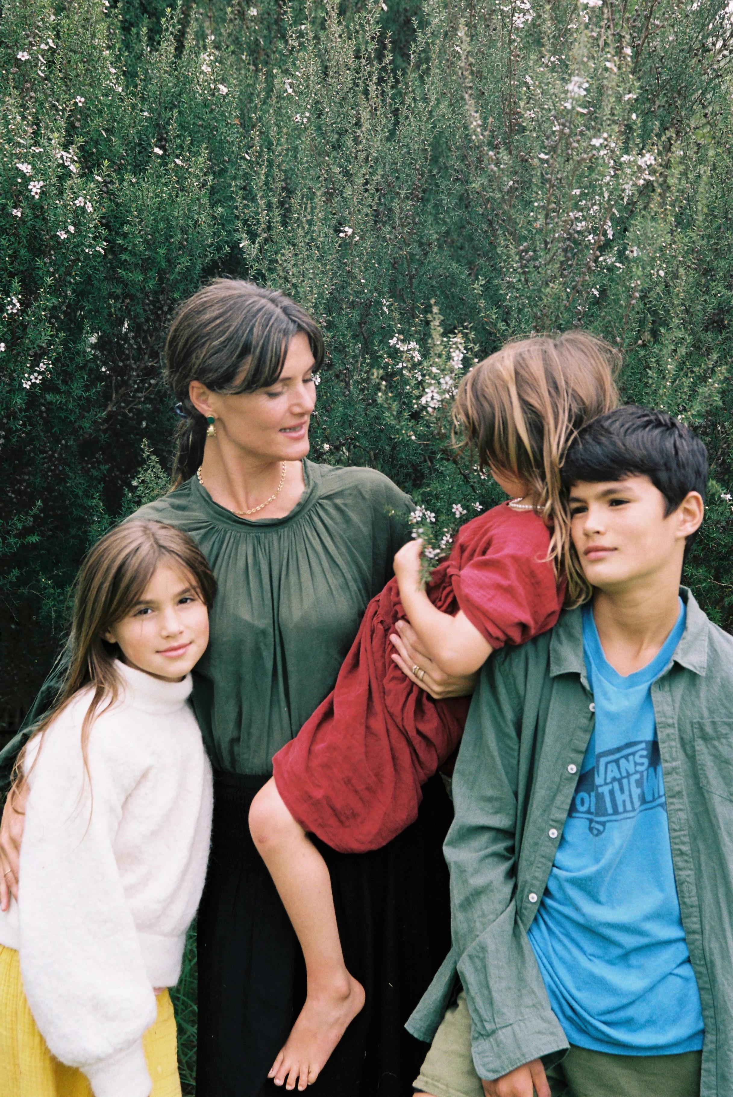 A woman stands with three children in front of dense greenery. She holds a child in red, with a girl in white to her left and a boy in blue to her right, both leaning towards her.