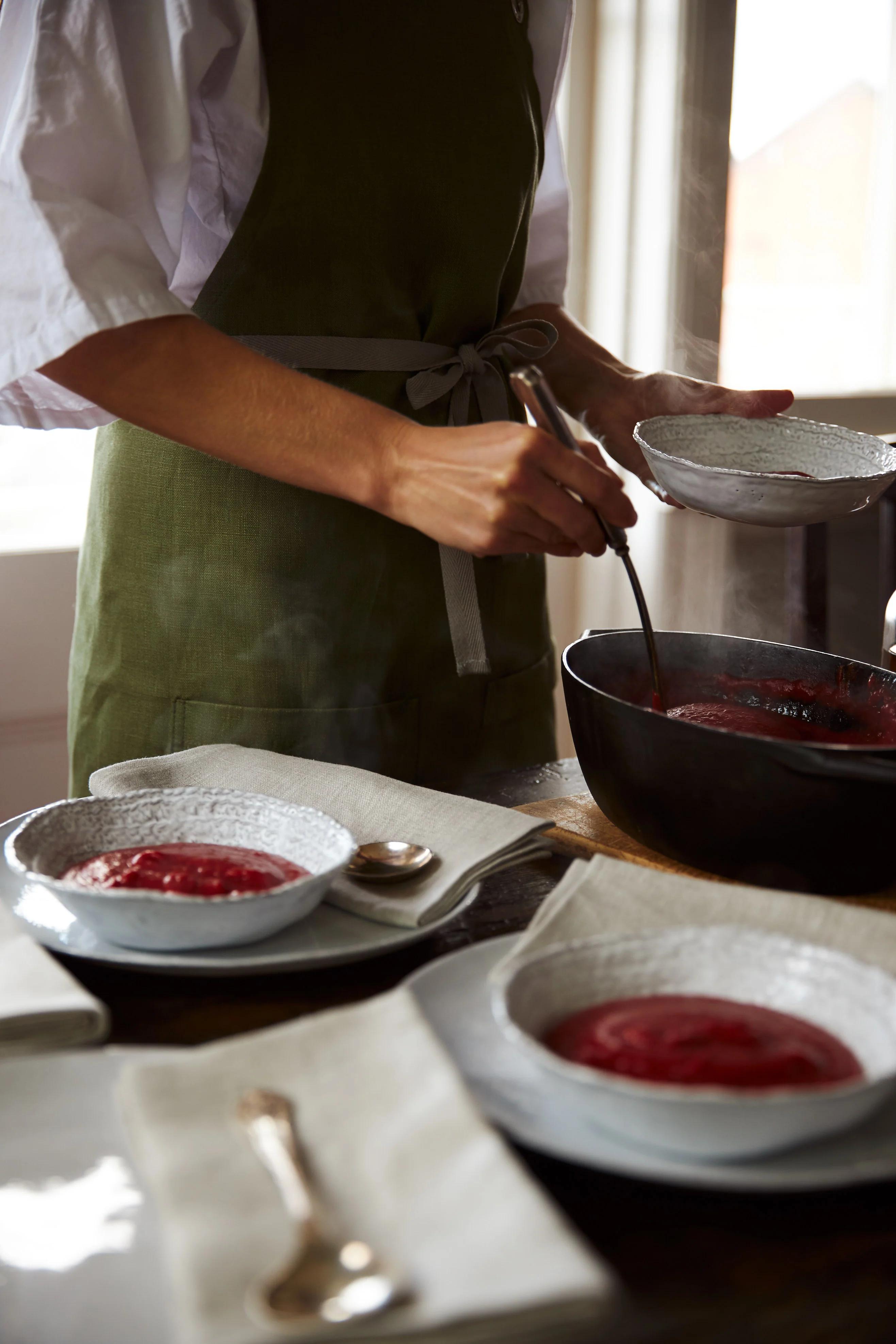 A person in a green apron serves hot beetroot soup from a black pot into white bowls. The table is set with napkins and is lit with sunlight from a background window.