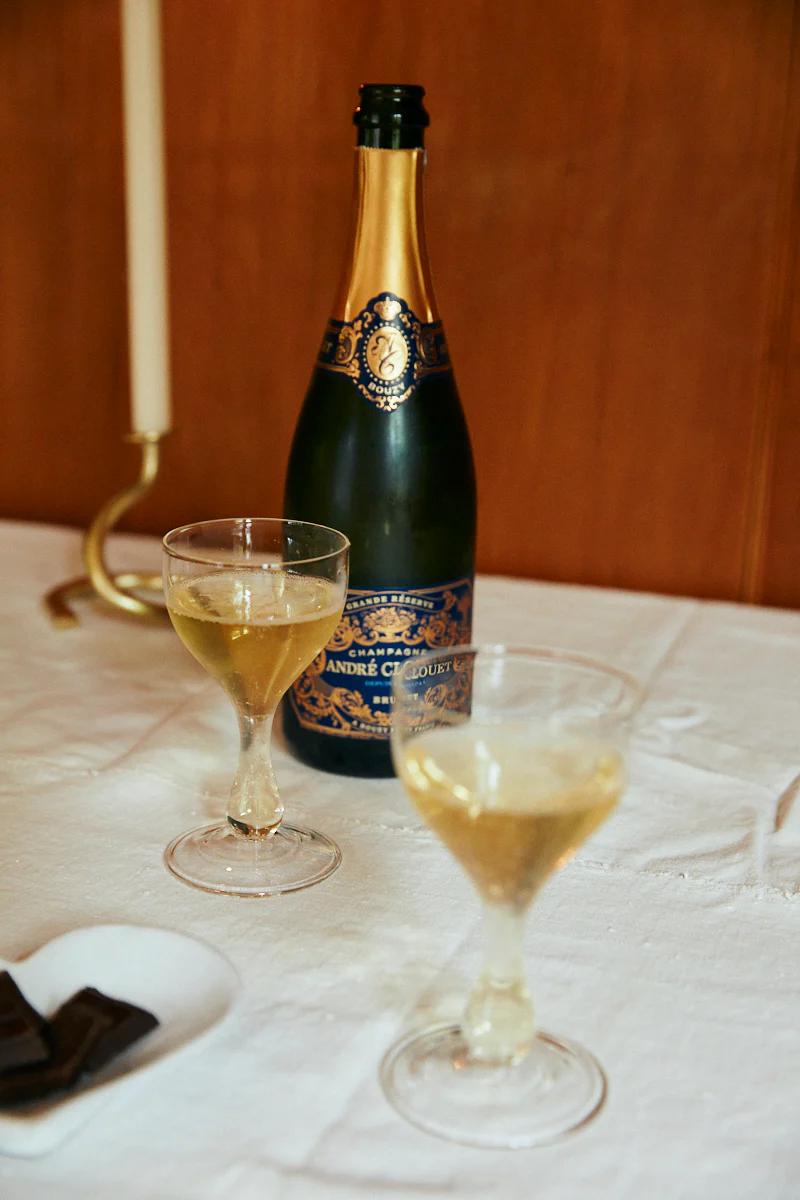 André Clouet Champagne bottle with gold and navy labels. Two filled wine glasses and dark chocolate pieces nearby.