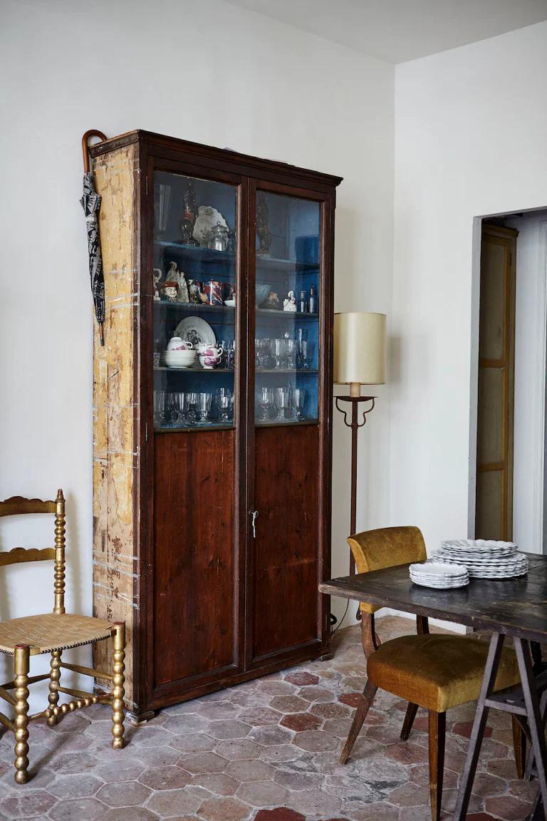 Parisian apartment room with antique wooden cabinet displaying Astier de Villatte glassware and ceramics. Wooden chair, floor lamp, and dining table with stacked dishes complete the cosy scene.