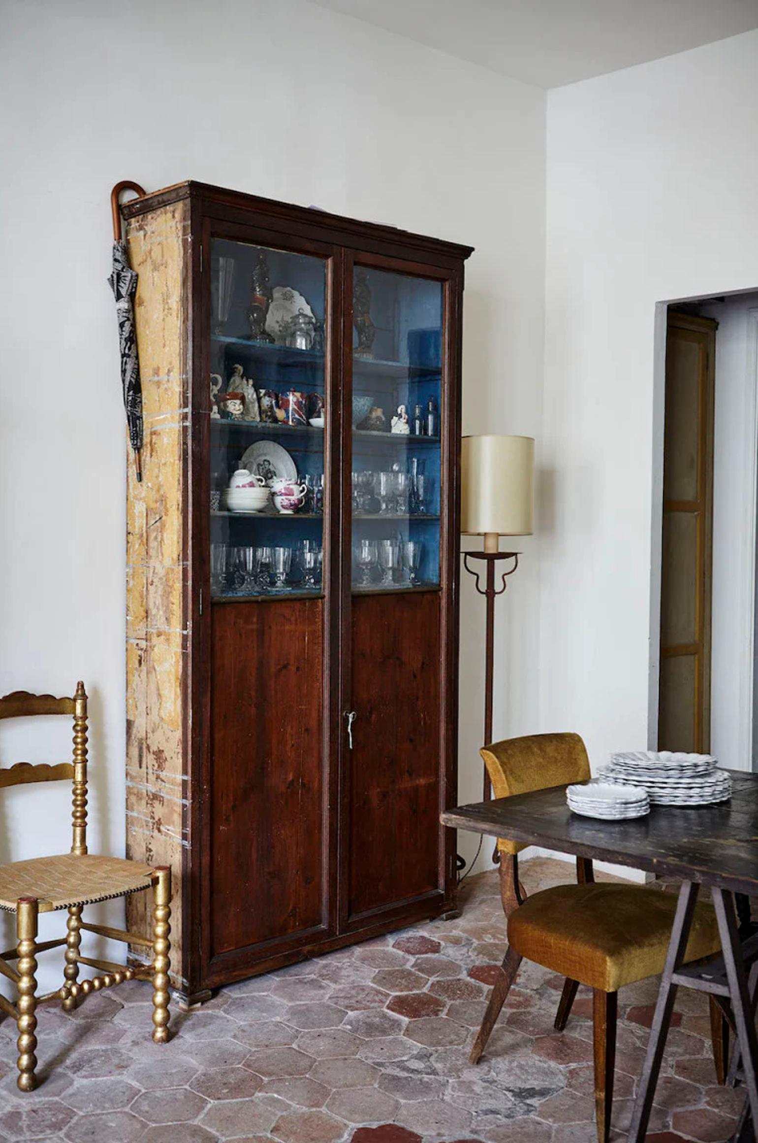 Parisian apartment room with antique wooden cabinet displaying Astier de Villatte glassware and ceramics. Wooden chair, floor lamp, and dining table with stacked dishes complete the cosy scene.