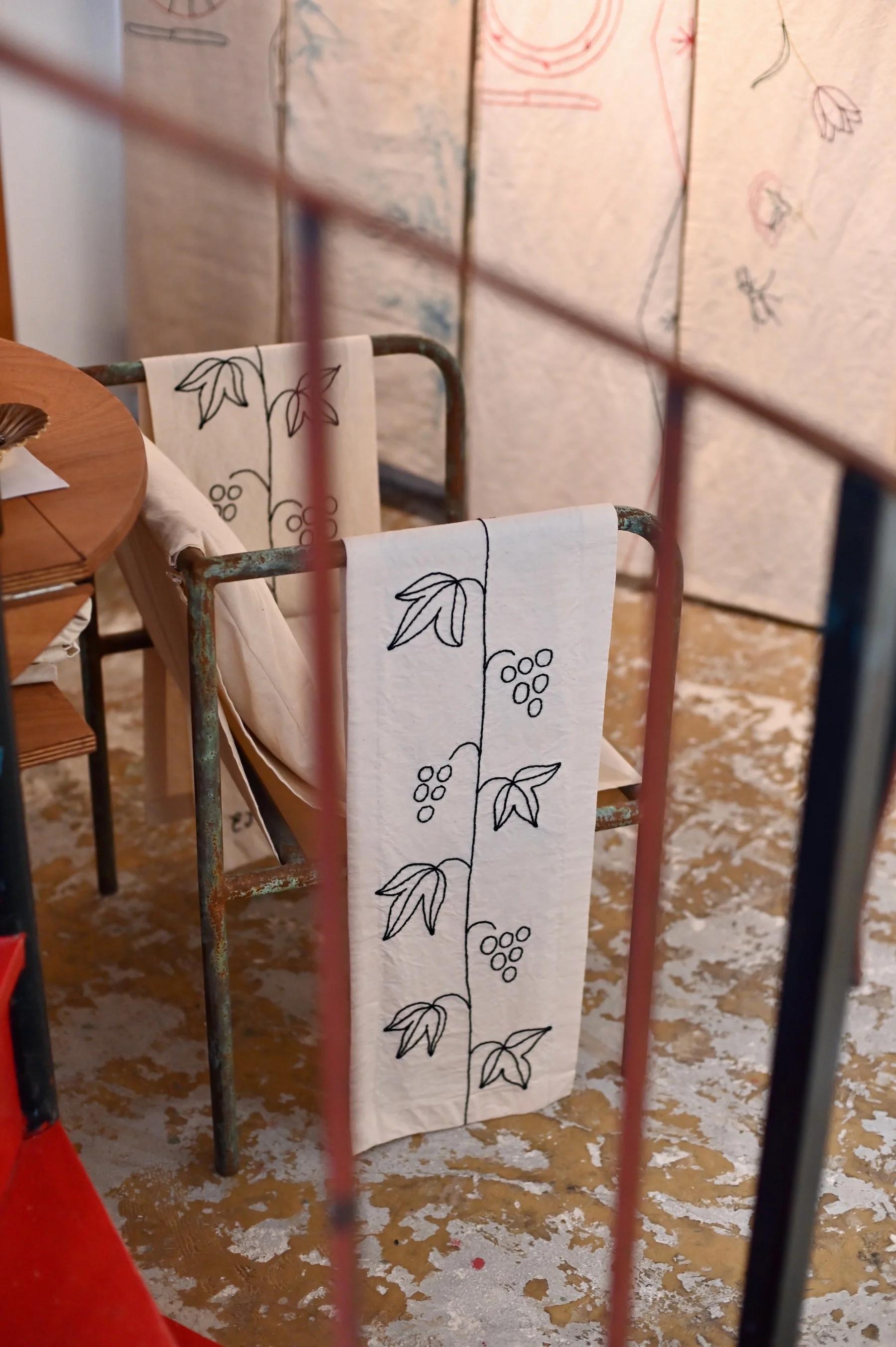 Metal-framed chair with white textile featuring hand-drawn vine pattern. Round wooden table nearby. Worn, painted floor.