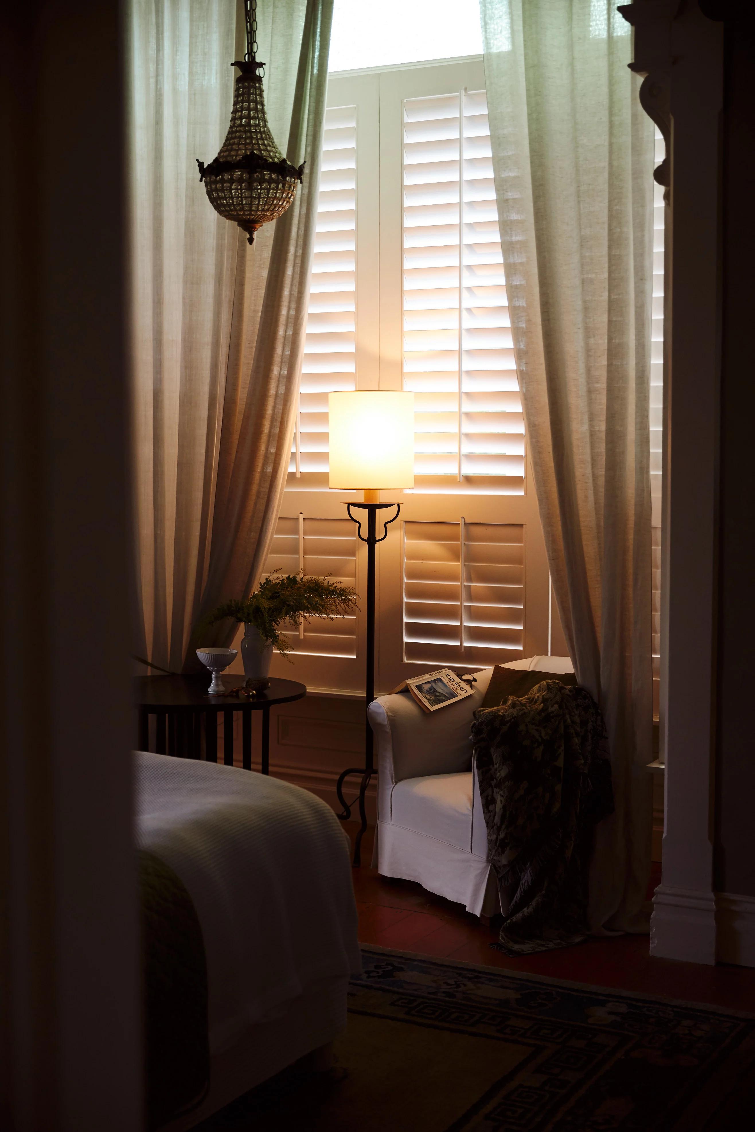 A cozy bedroom with a white armchair draped in a blanket, near tall windows with shutters and curtains. A side table holds a vase and magazine, illuminated by a floor lamp.