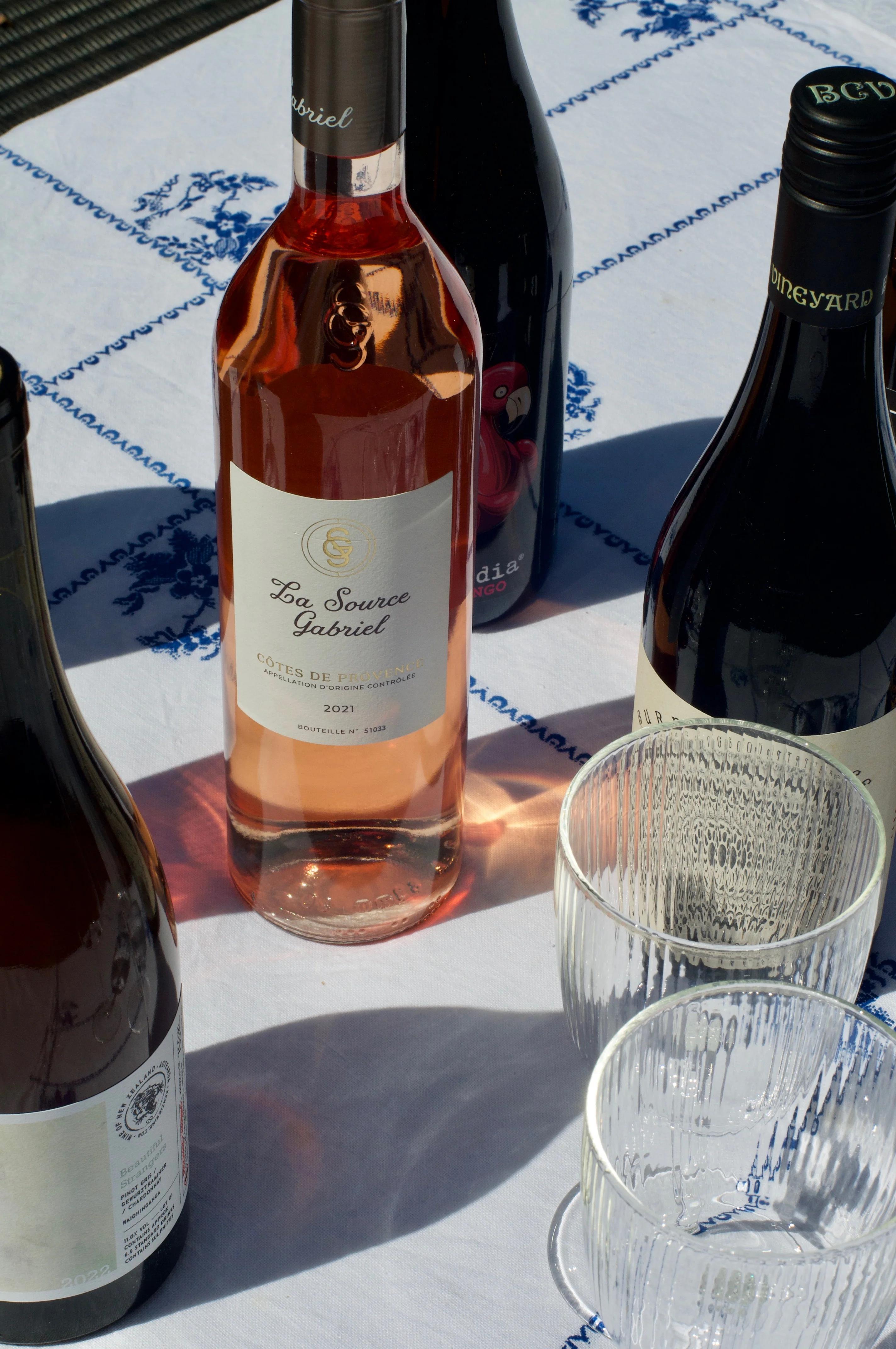 "La Source Gabriel" rosé bottle surrounded by other wines on patterned tablecloth. Two empty wine glasses beside bottles.