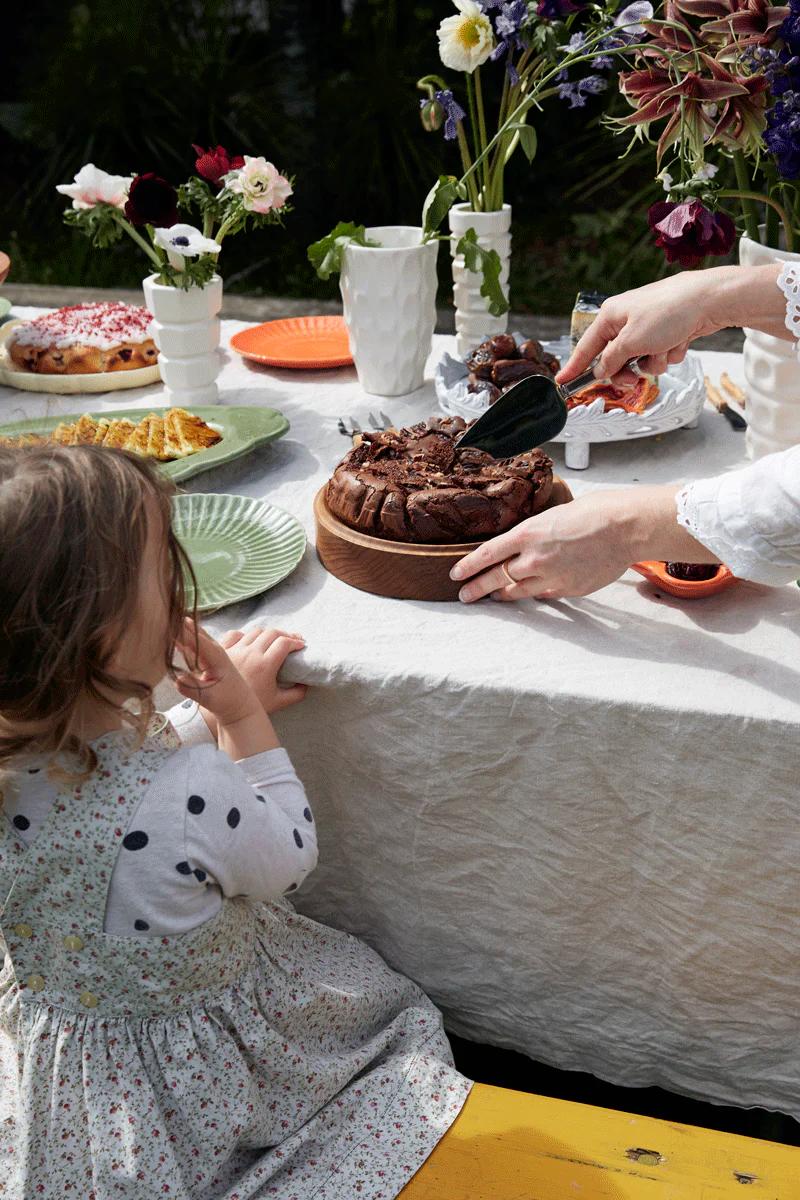 Child watches adult cutting chocolate bread on wooden board. Table covered with white cloth, plates of food, and flowers in vases. Child's sweet tooth anticipates baking aroma.