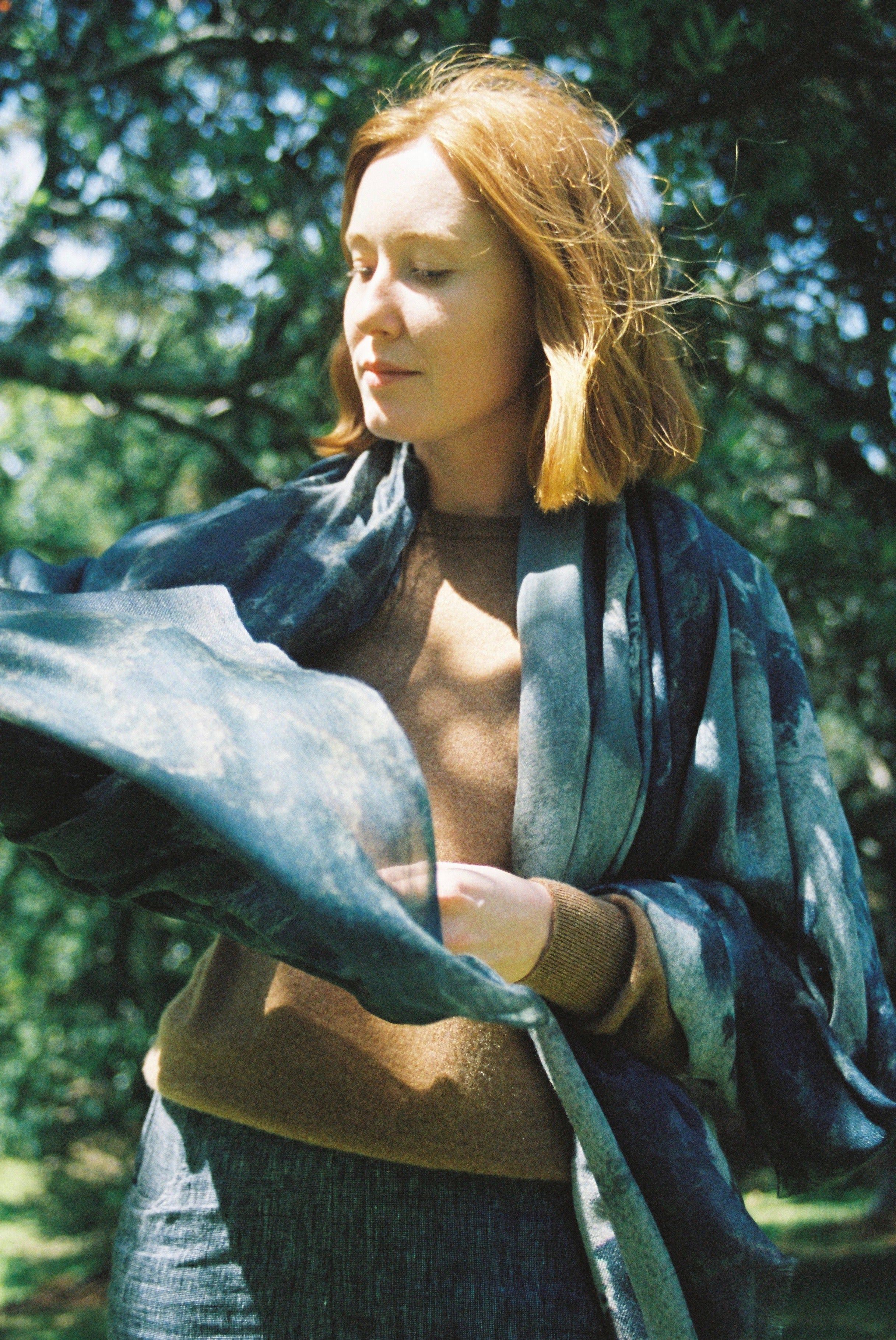 Light brown-haired person in brown sweater and large blue scarf. Looking down, surrounded by greenery and dappled sunlight.