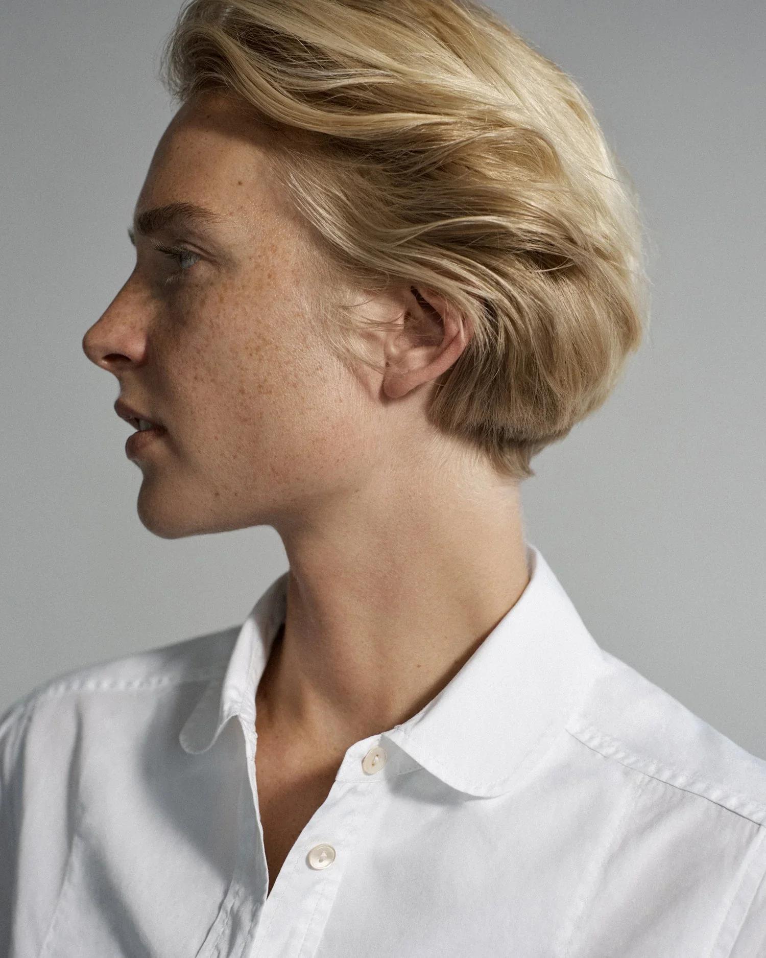 Profile of person with short blonde hair and freckles. White button-up shirt, neutral expression. Light gray background.