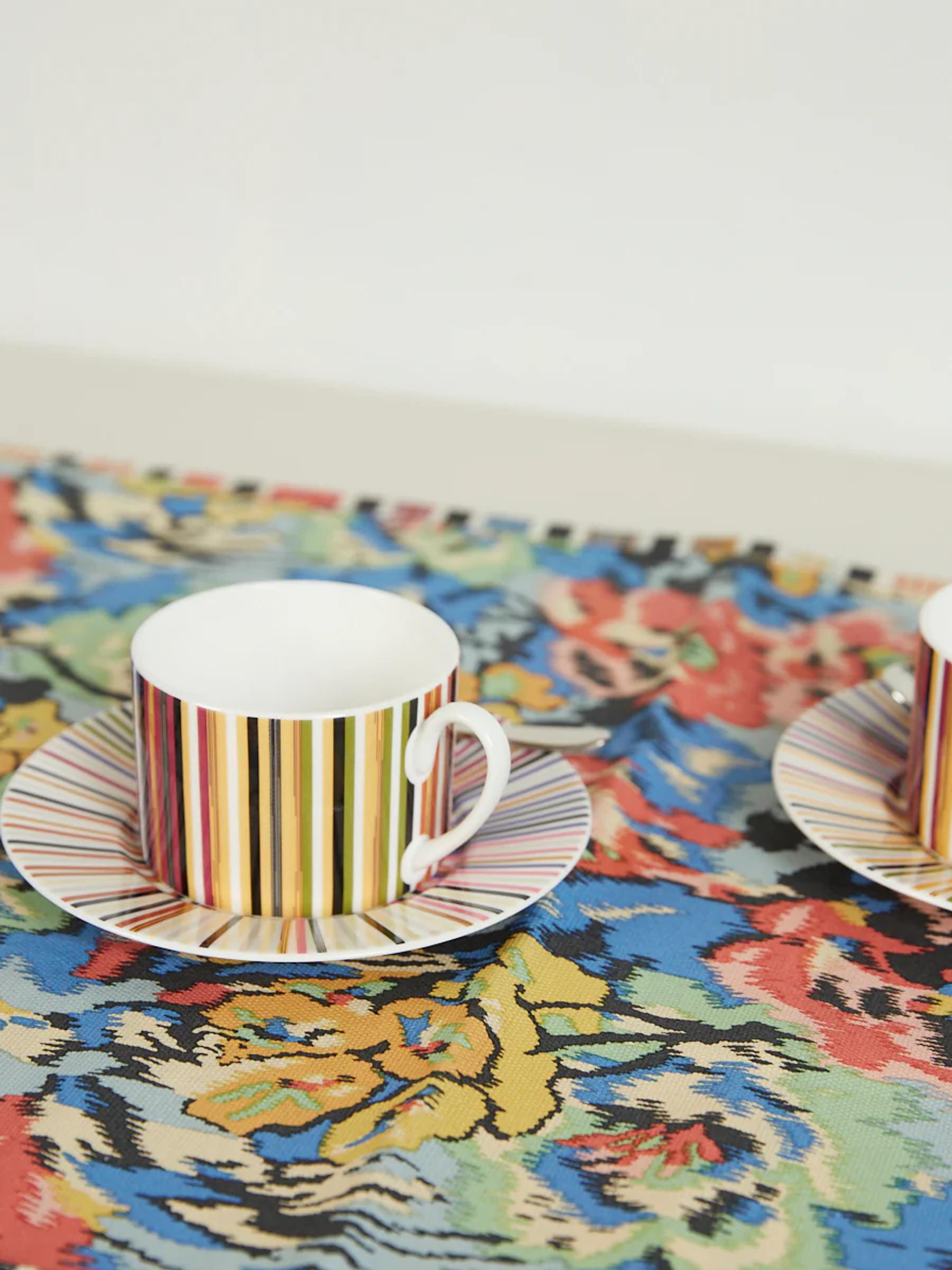 Two white teacups with colourful vertical stripes on matching saucers. Floral-patterned tablecloth. White background.
