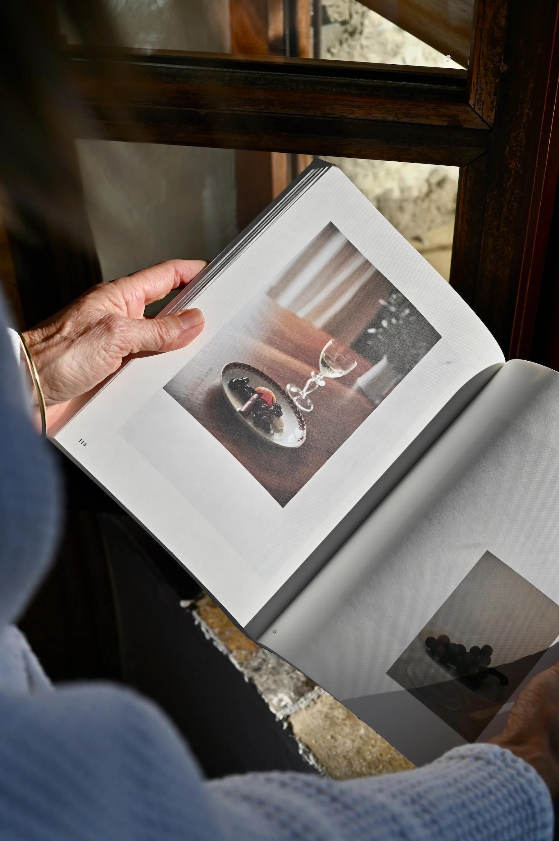 Person flipping through book with food photos. Held near sunlit wooden window frame.