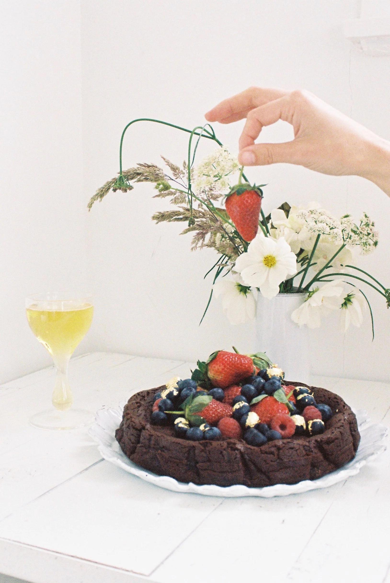 A hand holds a strawberry above a flourless chocolate cake with berries and gold details, set on a white plate with a glass of yellow liquid behind it.