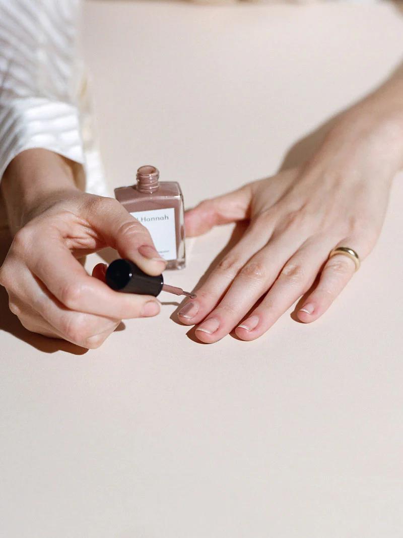 A person applies nude nail polish from J. Hannah while wearing a gold ring. The polish bottle is labeled “Hannah,” and the scene is set on a light-coloured surface.