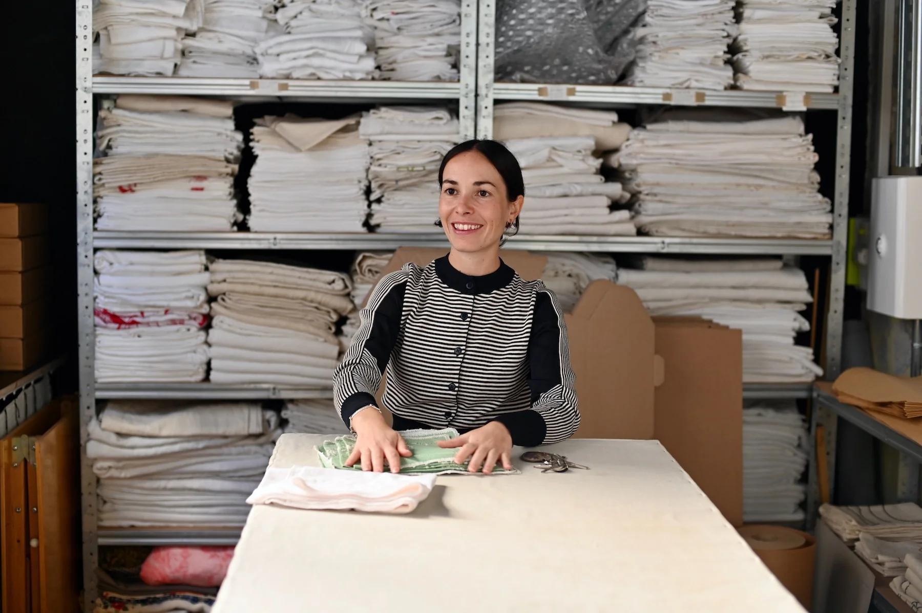 Smiling woman folds green fabric in well-organised storeroom. Shelves of white and beige textiles surround her.