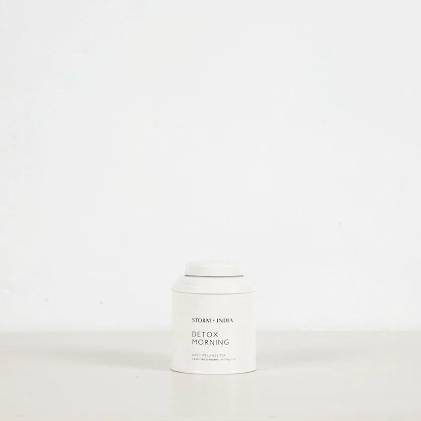 White cylindrical "Detox Morning" container from STORM + INDIA brand. Plain white background highlights minimalist design reflecting organic tea purity.