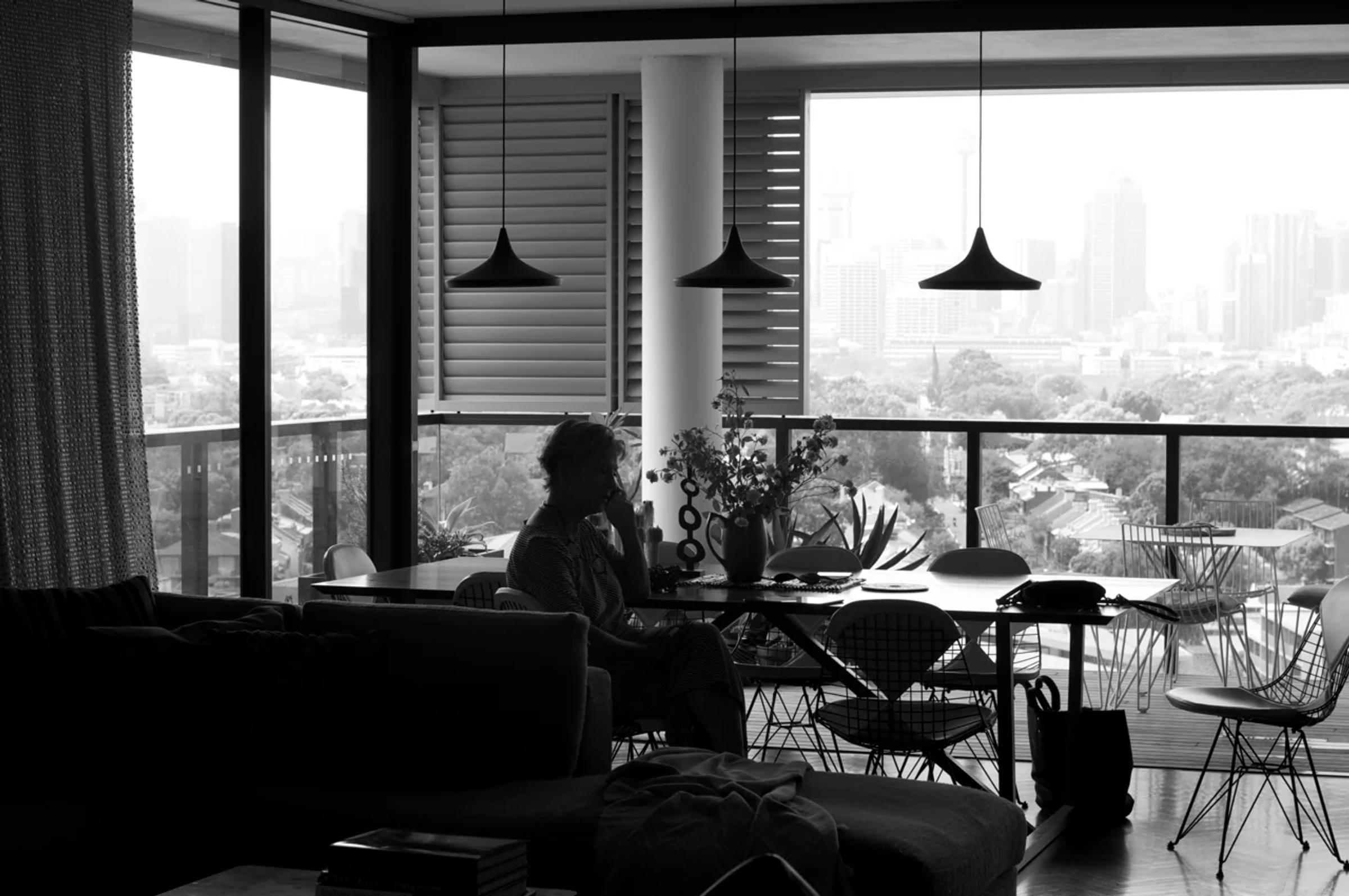 Black and white photo: Person at dining table in modern apartment. Sleek furniture and plants surround, with urban view through large windows.