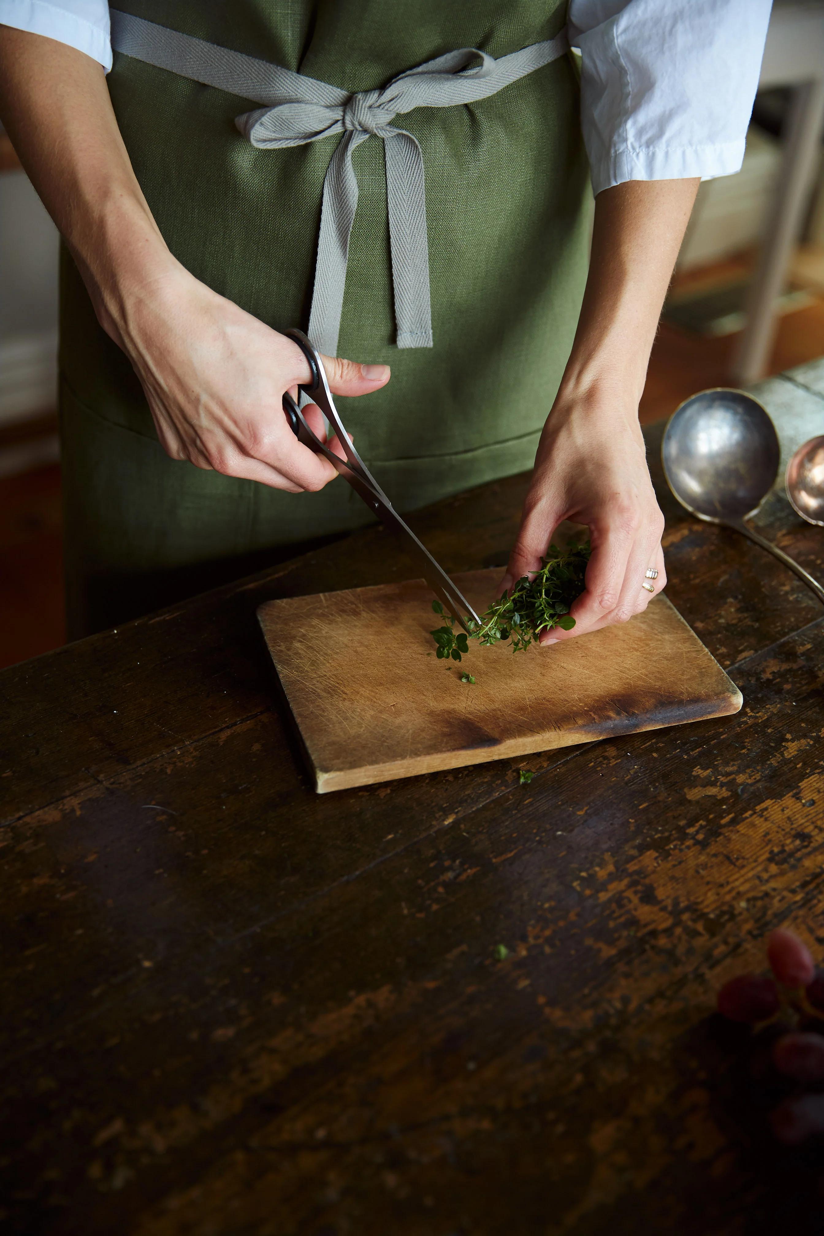 A person in a green apron cuts fresh herbs on a wooden board with scissors. The table is dark wood with scattered herbs, capturing preparation for a meal.