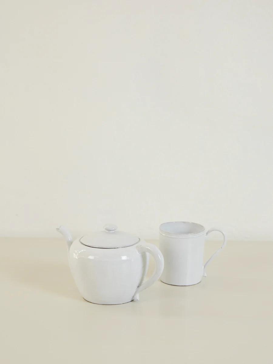 White ceramic teapot and mug on light surface. Plain, off-white background.