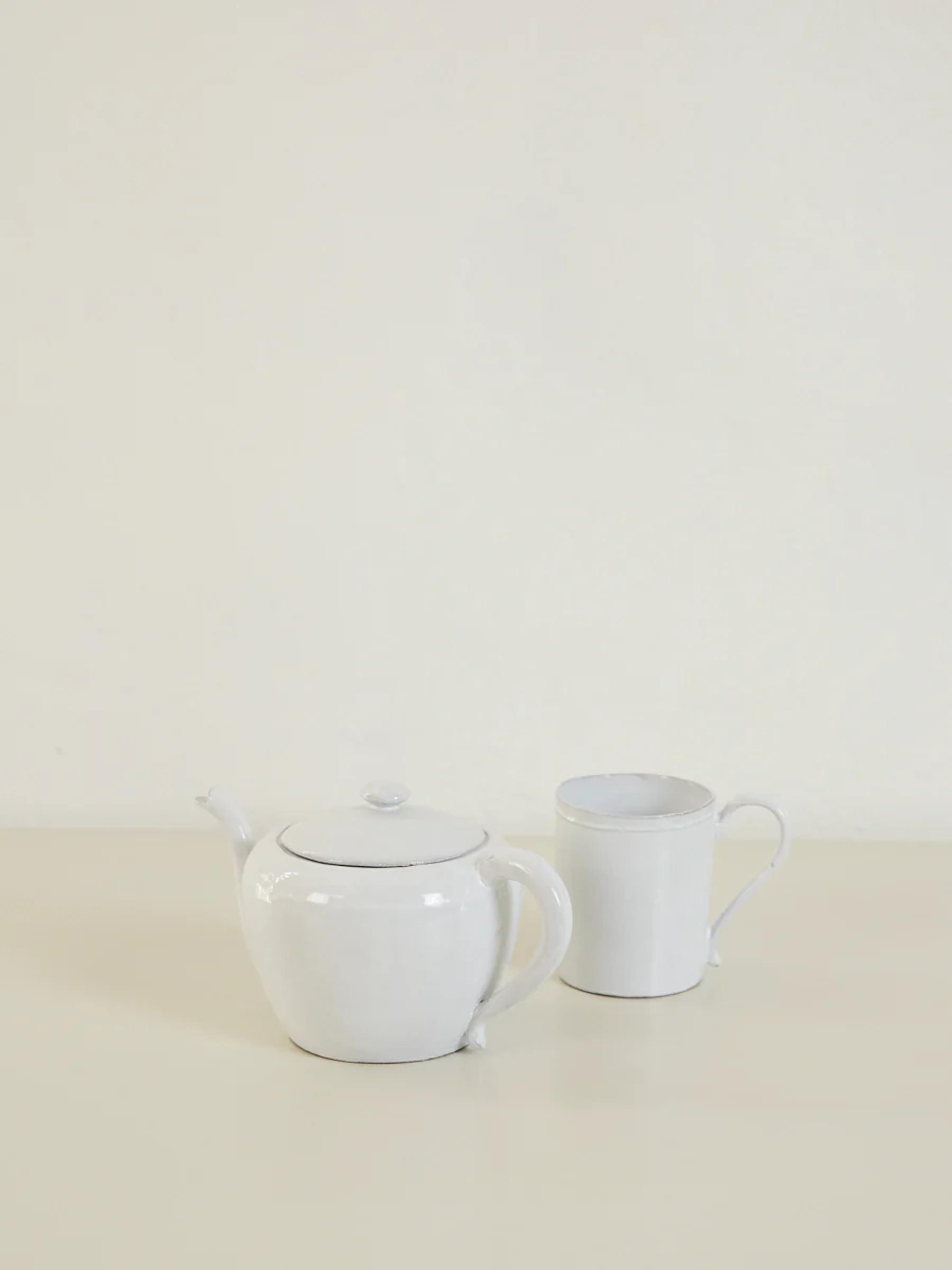 White ceramic teapot and mug on light surface. Plain, off-white background.