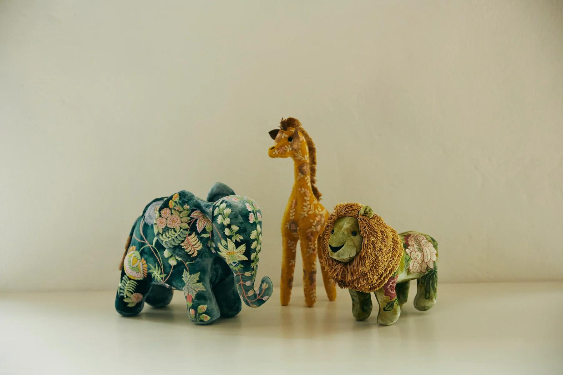 Three stuffed animals - an elephant, giraffe, and lion - sit on a flat surface. Each features vibrant, embroidered fabric patterns with floral and leafy designs in contrasting colours.