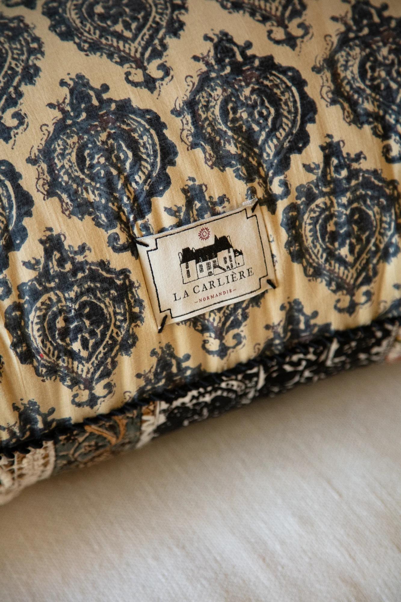 Close-up of vintage-style cushion. Intricate black designs on beige. Label reads "L. Carlier - Versailles" with house logo.