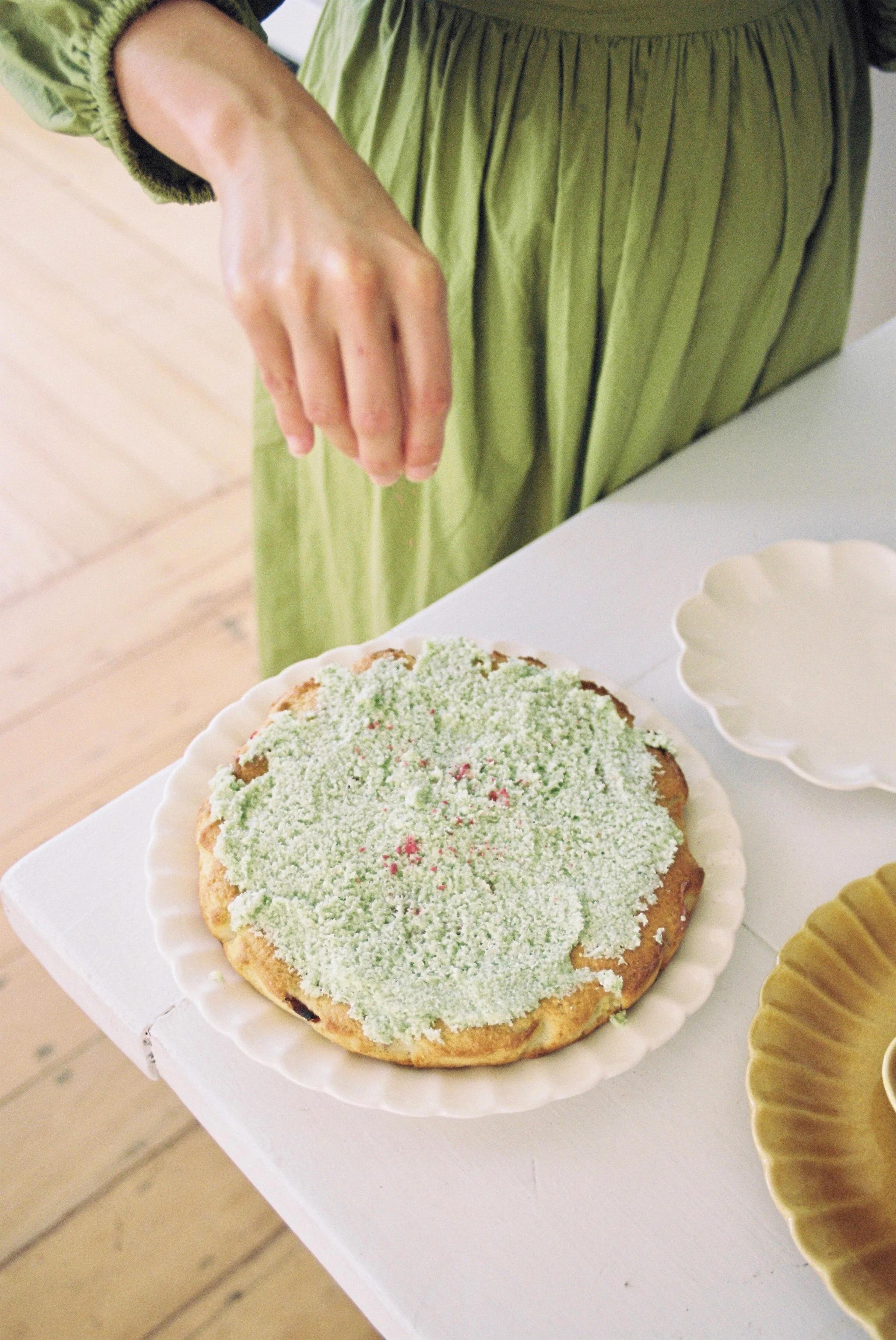 Person in green dress sprinkling toppings on green-frosted pie. White plate on white table.