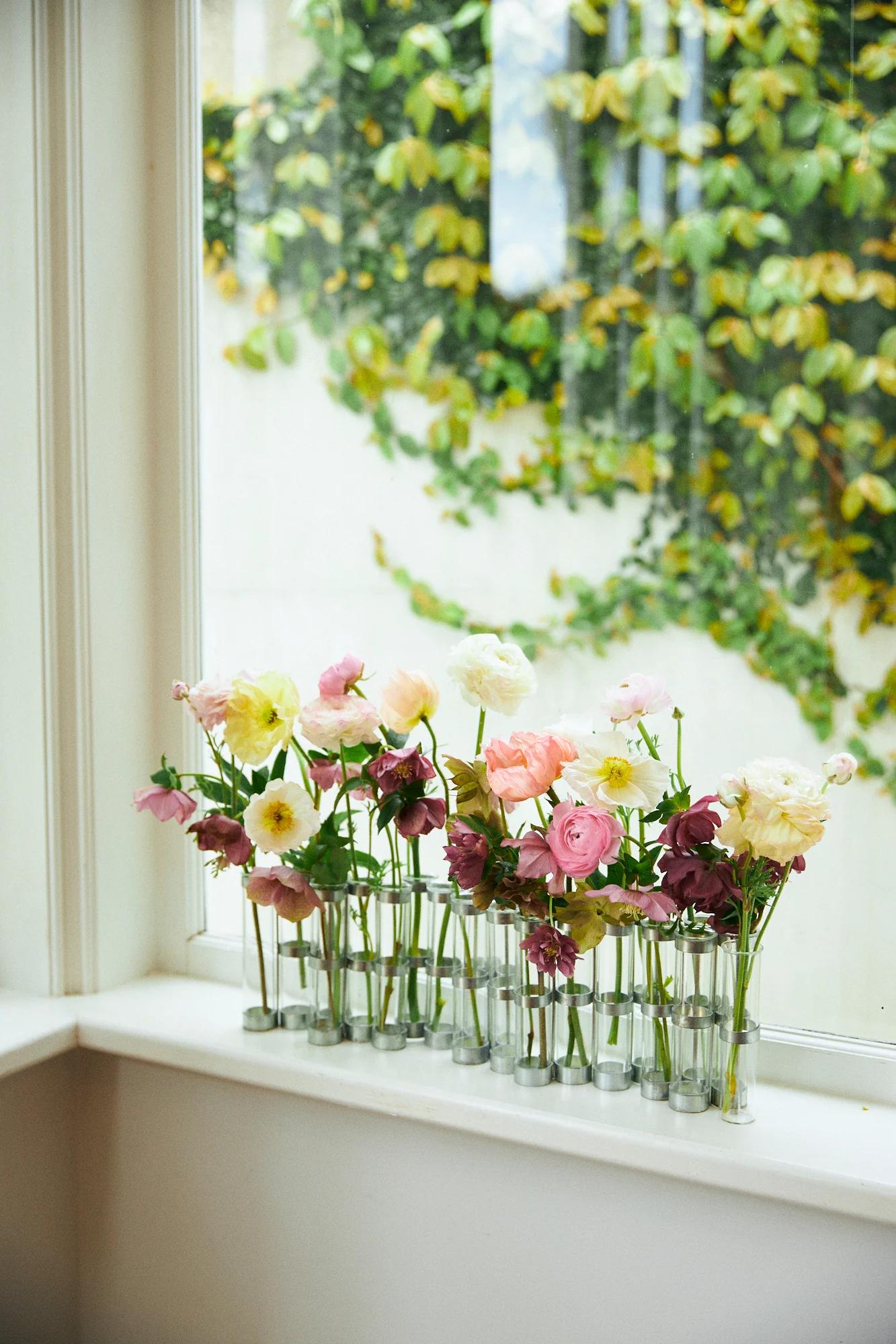 Glass vases with white, pink, and purple flowers on windowsill. Window shows ivy-covered outdoor wall.