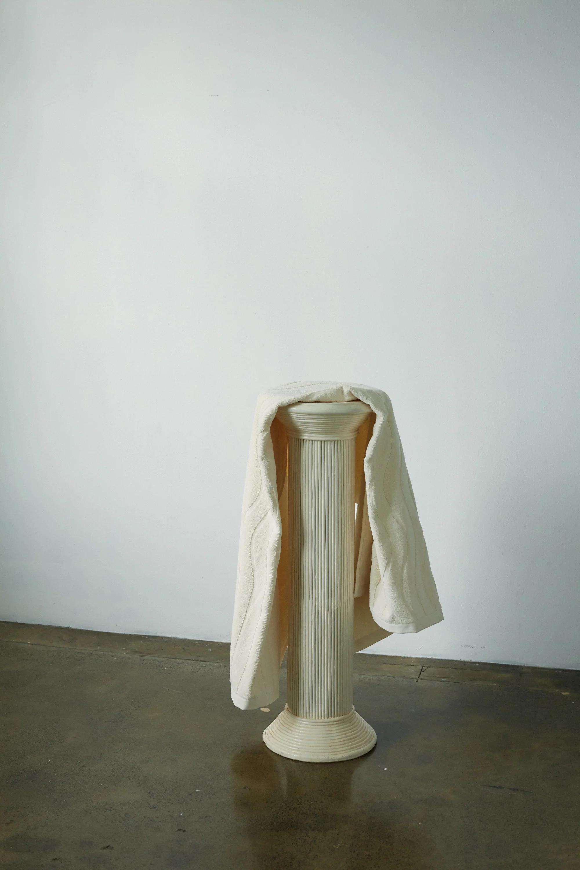 A white fluted column pedestal stands on a polished concrete floor. A cream-colored fabric drapes over the top, cascading down the sides, showcasing Baina’s modern, minimalistic aesthetic.