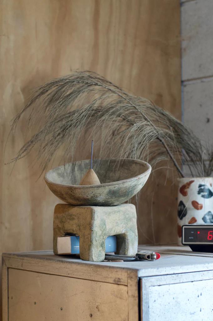 Ceramic bowl on stone stand with burning incense, dried grasses nearby. Lighter and digital clock on concrete shelf. Background wall mimics NZ wood textures.