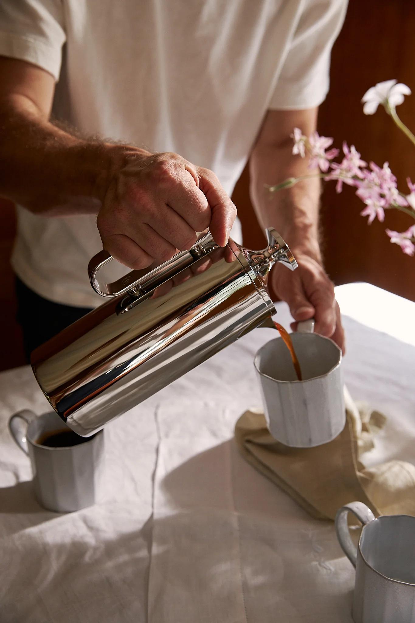 Man pours coffee for a friend, seen through purple flowers 