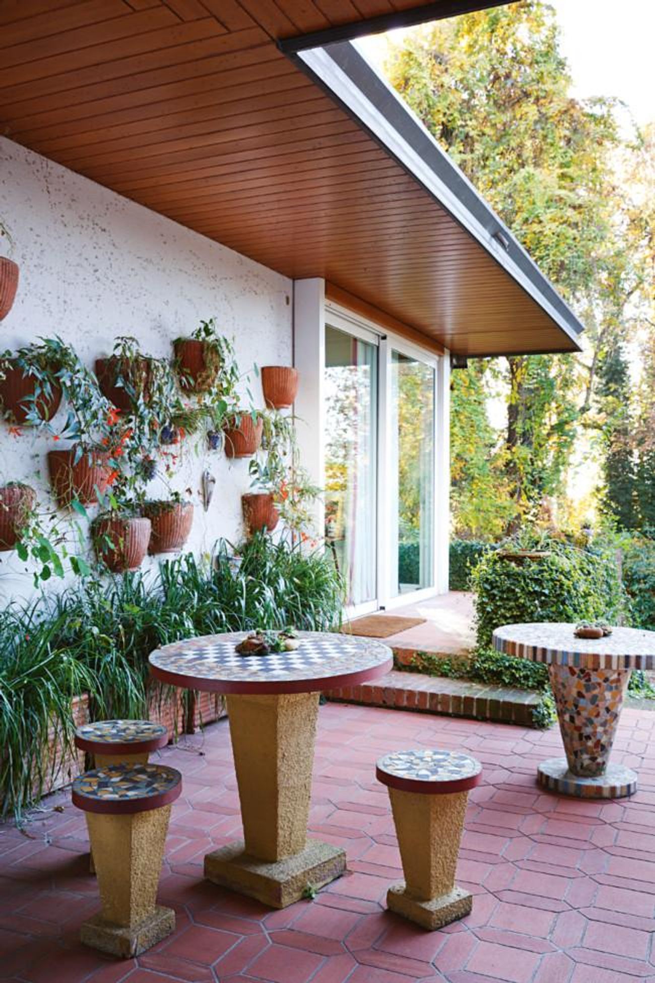 Outdoor patio with tiled table and stools. Potted plants on white wall. Lush greenery surrounds. Wooden ceiling contrasts with plants.