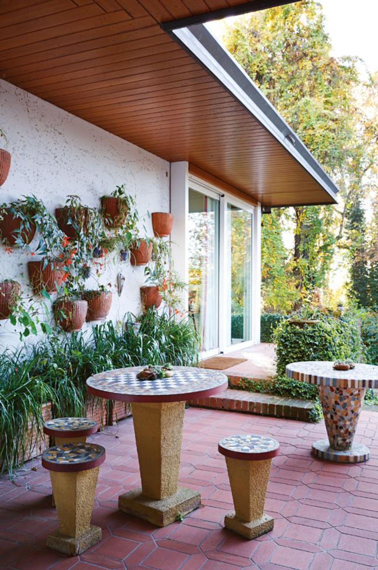 Outdoor patio with tiled table and stools. Potted plants on white wall. Lush greenery surrounds. Wooden ceiling contrasts with plants.