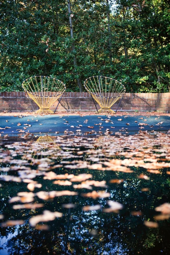 Two yellow wire chairs on patio. Reflective surface with fallen leaves. Brick wall and dense foliage in background.