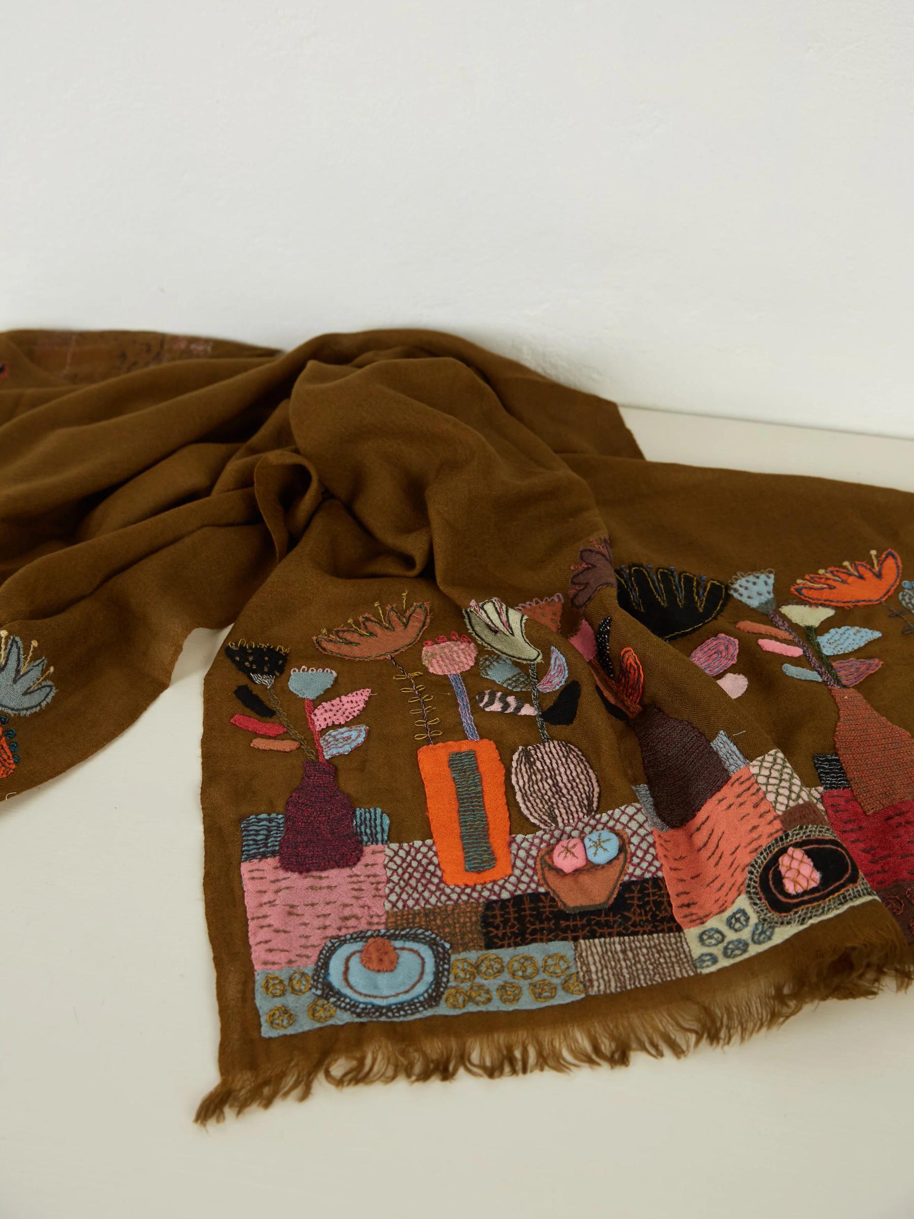 A brown scarf by Sophie Digard with vibrant embroidered abstract plant designs in pink, red, orange, green, and blue, and fringe detail.