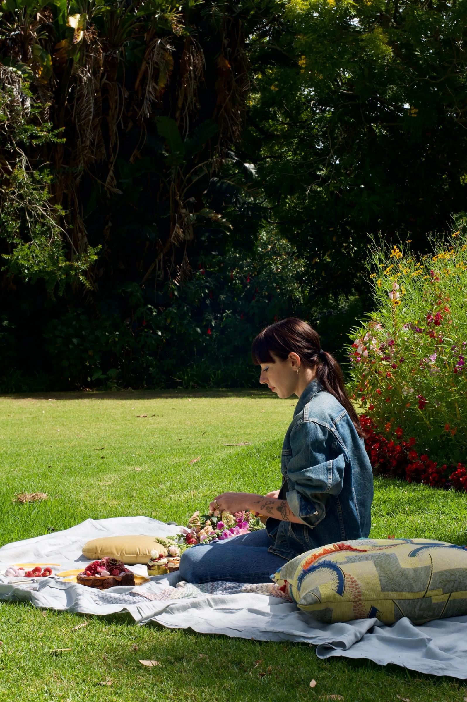 A person with a ponytail arranges flowers on a picnic blanket surrounded by greenery, with fruits, snacks, and a colourful cushion from The Caker bakery’s collection.