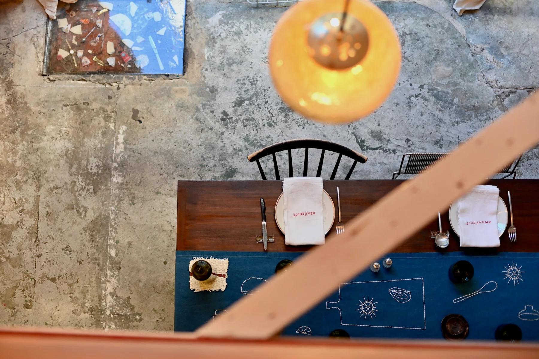 Top-down view of rustic table set for two. Blue table runner on weathered concrete floor. Warm light fixture above.