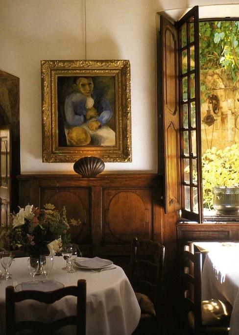 Cosy restaurant interior with wooden paneling. Round table, floral centerpiece, framed painting. Open window shows Provencal greenery.